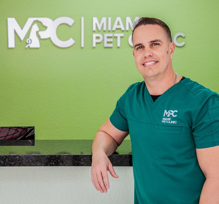 A person in a teal uniform stands smiling beside a counter in front of the "Miami Pet Clinic" sign.