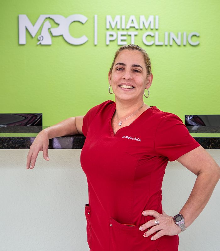 A smiling person in a red uniform stands confidently in front of a green wall at the Miami Pet Clinic.