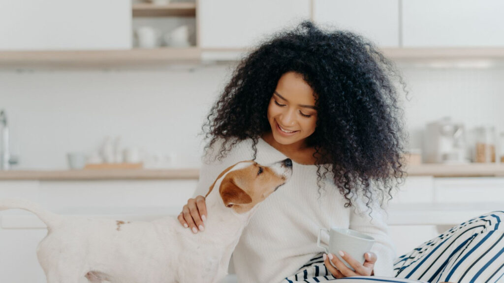 A woman with curly hair smiles at a dog while holding a mug in a kitchen setting.