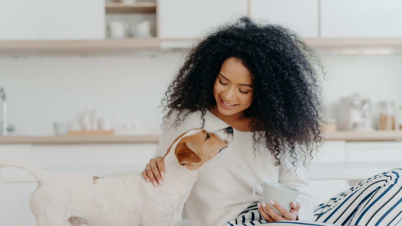 A woman with curly hair smiles at a dog while holding a mug in a kitchen setting.